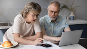 An older couple looking a spreadsheets and calculator in front of a computer